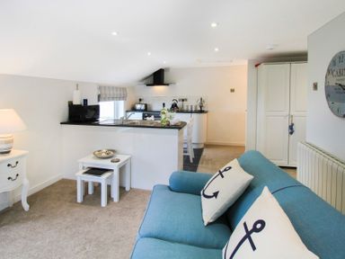 A living area with a blue sofa and anchor cushions next to a small white nested table and a kitchen with a black countertop at Amolca in Llaneilian