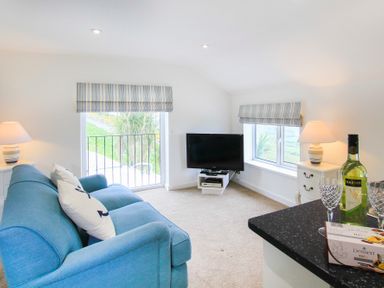 A living room with a blue sofa two windows with striped blinds a television on a stand two lamps a bottle of wine glasses and a box of chocolates at Amolca in Llaneilian