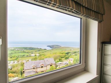 A window with a striped blind showing a coastal landscape with houses green fields and the sea at Amolca in Llaneilian