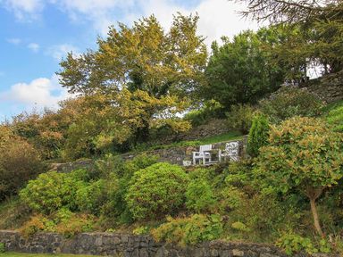 A garden area with stone walls and white chairs surrounded by trees and shrubs at Amolca in Llaneilian