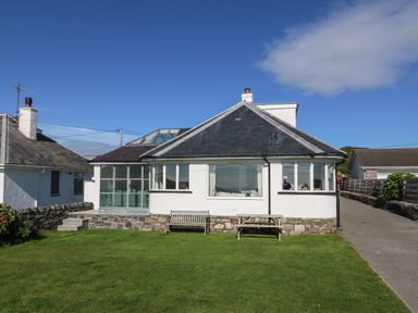 A single-story house with white walls and a dark roof a wooden bench and table on the lawn at Angorfa - Trearddur Bay