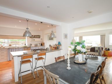 An open kitchen and dining area with a kitchen island and high chairs and a wooden dining table with flowers at Angorfa - Trearddur Bay