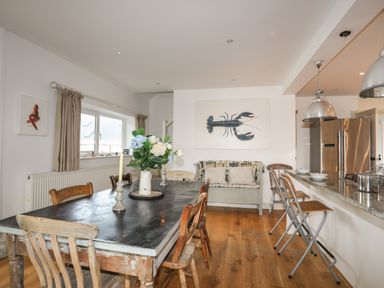A dining area with a large table and chairs a sofa with cushions lobster artwork on the wall and a kitchen counter with stools at Angorfa - Trearddur Bay in Trearddur Bay
