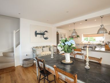 A dining area with a dark table and wooden chairs a vase with flowers on the table candles on the table a kitchen with pendant lights and a lobster artwork on the wall at Angorfa - Trearddur Bay