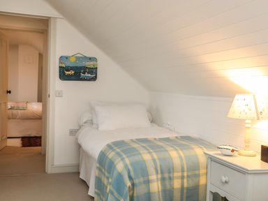 A bedroom with a single bed covered with a plaid blanket a bedside table with a lamp and an open door showing another bed at Angorfa - Trearddur Bay in Trearddur Bay