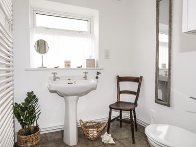 A bathroom with a pedestal sink a wooden chair a wicker basket and a tall mirror at Angorfa - Trearddur Bay in Trearddur Bay