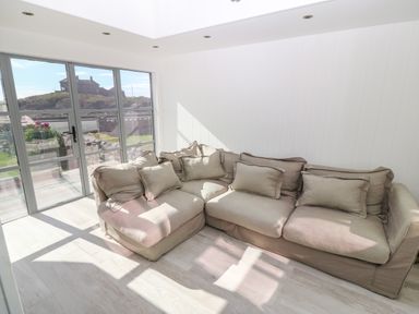 A corner sofa with multiple cushions in a room with glass doors and a view of houses and a road at Angorfa Trearddur Bay