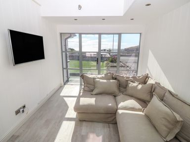 A seating area with a beige sectional sofa and a wall-mounted TV near glass doors with a view of the outdoors at Angorfa Trearddur Bay