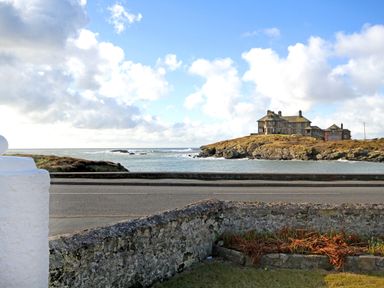 A coastal view with a stone wall in the foreground a road and a stone building on a rocky hill at Angorfa - Trearddur Bay in Trearddur Bay