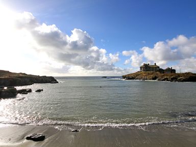 A coastal scene with a sandy beach, rocky shoreline, and a house on a small peninsula under a cloudy sky at Angorfa - Trearddur Bay in Trearddur Bay