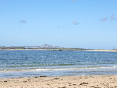 A sandy beach with seaweed and waves with a body of water and hills in the background at Arkoo in Rhosneigr