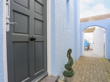 A blue exterior wall with a black door and a potted topiary near an open white garden gate with a table and chairs visible beyond at Arkoo in Rhosneigr