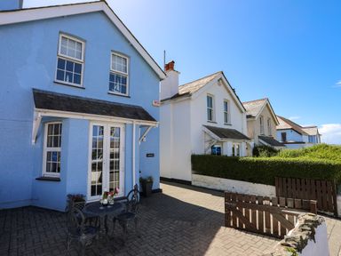 A blue house with a small outdoor table and chairs on a paved patio next to white houses and hedges at Arkoo in Rhosneigr