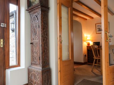 A wooden carved grandfather clock next to a glass door leading to a room with a wooden chair a desk and a lamp at Barras Cottage in Brynsiencyn