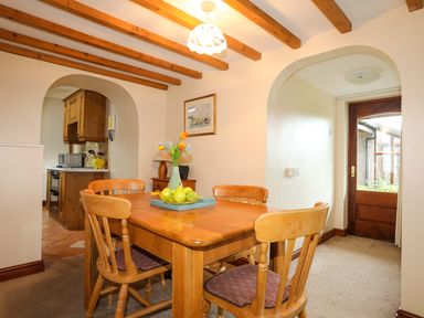 A dining room with a wooden table and chairs next to an arched kitchen doorway and a wooden door at Barras Cottage in Brynsiencyn
