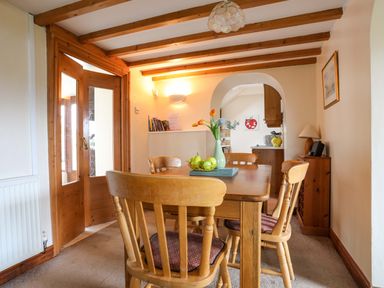A dining room with a wooden table and chairs a vase with flowers and a bowl of fruit on the table at Barras Cottage in Brynsiencyn