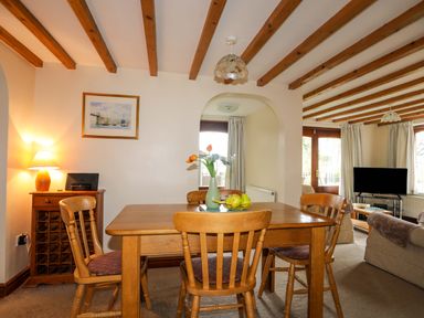 A dining room with wooden table and chairs next to a living room with sofa and television at Barras Cottage in Brynsiencyn