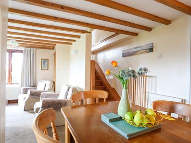 A dining table with a vase of flowers and a fruit bowl in a room with armchairs and wooden ceiling beams at Barras Cottage in Brynsiencyn