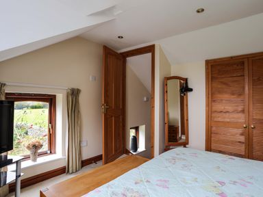 A bedroom with a bed, wooden wardrobe, mirror, and window looking outside at Barras Cottage in Brynsiencyn