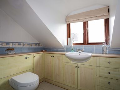 A bathroom with a wall-mounted sink a toilet under a slanted ceiling light wood cabinets and a window with beige blinds at Barras Cottage Brynsiencyn