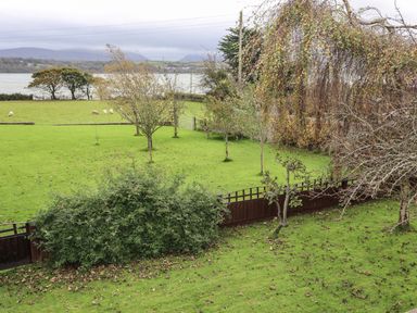 A grassy yard with trees and a wooden fence overlooking sheep and water at Barras Cottage in Brynsiencyn