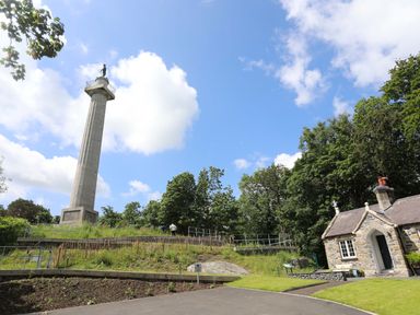 A tall monument with a statue on top near a small stone building surrounded by trees at Barras Cottage in Brynsiencyn