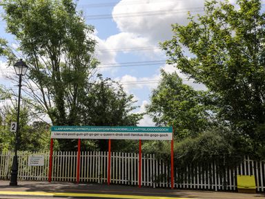 A railway platform with a sign displaying a long place name and trees behind a white fence at Barras Cottage in Brynsiencyn