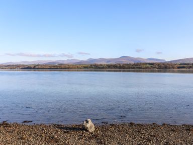A rocky shore with a body of water and hills in the background at Barras Cottage in Brynsiencyn