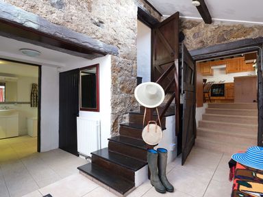 An entryway with stone walls showing stairs leading to a kitchen a black door a bathroom and a hat stand with boots and hats at Foel Stable Cottage Brynsiencyn