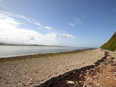 A rocky beach with water and a green hill on the right side at Foel Stable Cottage in Brynsiencyn