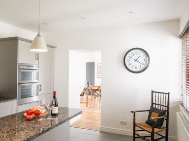 A kitchen with a granite countertop holding wine, glasses, bread, tomatoes, and an onion with a wooden chair and a clock on the wall at Bryn Deryn in Criccieth