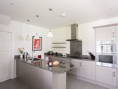 A kitchen with a granite countertop island, pendant lights, built-in ovens, cabinets, and a wall with shelves holding glasses at Bryn Deryn in Criccieth
