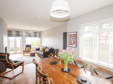 A dining table with flowers and wooden chairs next to a living area with sofa and armchairs at Bryn Deryn in Criccieth