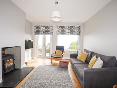 A living room with a gray sofa with yellow and white pillows a black armchair with a yellow pillow a patterned pouf a fireplace and a wall-mounted television at Bryn Deryn in Criccieth
