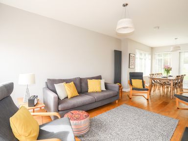 A living room with gray sofa and armchairs with yellow cushions a small wooden table with a lamp an ottoman and a dining table with chairs and flowers at Bryn Deryn in Criccieth