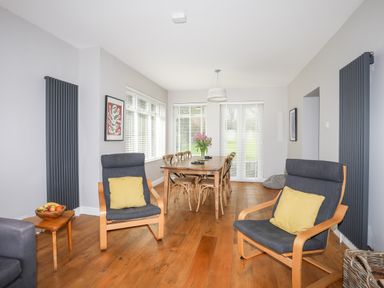 A dining room with a wooden table and chairs two armchairs with yellow cushions a small side table with a fruit bowl and large windows at Bryn Deryn in Criccieth