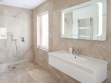 A bathroom with a walk-in shower a window with blinds a wall-mounted sink and an illuminated mirror at Bryn Deryn in Criccieth