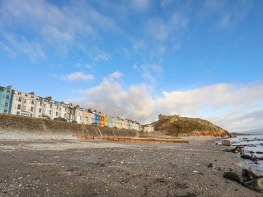 A pebble beach with colorful houses and a hill with a castle in the background at Bryn Deryn Criccieth