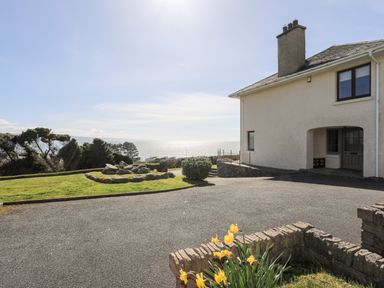An exterior view of a house with a driveway garden bed with yellow flowers and a grassy area overlooking the sea at Bryn Deryn in Criccieth