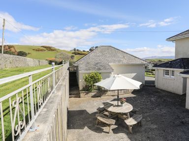 A patio area with a round wooden picnic table and white umbrella near a building with a sloped roof and a grass lawn bordered by a stone wall at Bryn Deryn in Criccieth