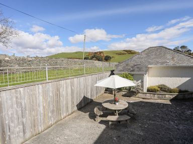 An outdoor patio area with a round wooden table, benches, and a white umbrella surrounded by a stone wall and wooden fence at Bryn Deryn in Criccieth