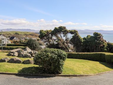A garden with shrubs rocks and grass overlooking houses and a sea with hills in the background at Bryn Deryn in Criccieth