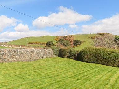 A lawn with trimmed hedges and a stone wall with green hills and blue sky in the background at Bryn Deryn in Criccieth