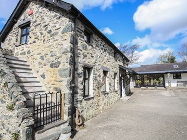 An outdoor view of a stone building with stairs and a gate next to a paved area with plants at Bryn Eira Stables in Llanfair PG