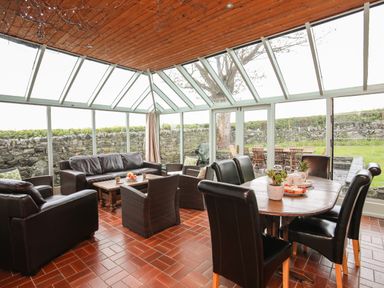A conservatory with black leather sofas a wooden dining table and chairs and glass walls at Bryn Eira Stables Llanfair PG