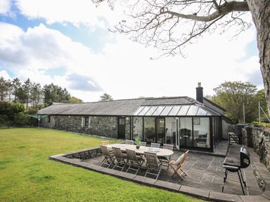 A stone house with a glass extension and outdoor dining table on a patio with chairs and a barbecue at Bryn Eira Stables in Llanfair PG