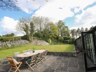 An outdoor patio with a wooden dining table and chairs next to a grass lawn and trees at Bryn Eira Stables in Llanfair PG