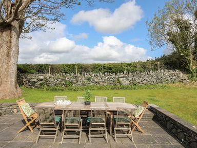 An outdoor patio area with a wooden table and chairs on a stone floor with a stone wall and grass behind at Bryn Eira Stables in Llanfair PG
