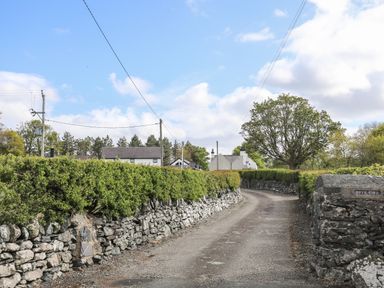 A curved paved driveway with stone walls and green hedges leading to houses with trees and power lines at Bryn Eira Stables in Llanfair PG