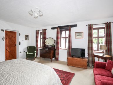 A bedroom with a bed a green armchair a wooden dresser with a mirror a television on a wooden chest red and plaid curtains and a red sofa at Bryn Eira Stables in Llanfair PG